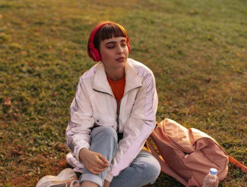 Charming short-haired woman in jeans and white jacket sits on grass. Cool girl in stylish outfit listens to music in red headphones outdoors.