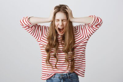 Girl screaming from mental instability. Portrait of depressed fed up young woman shouting and holding hands on head, closing eyes, being stressed and miserable, standing over gray wall devastated. Emotions concept